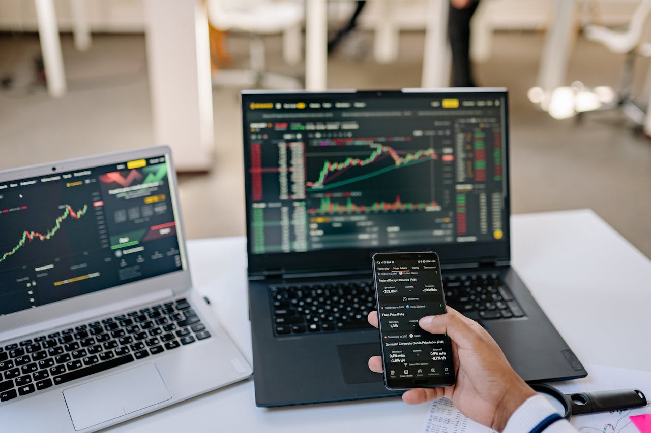 A professional tracks stock market trends using two laptops and a smartphone, showcasing digital finance tools.